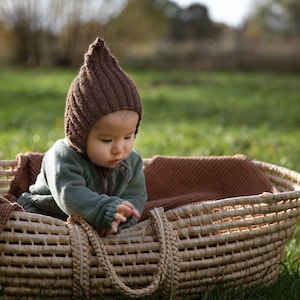 Könnte beinhalten: Ein Baby mit einer braunen Strickmütze sitzt in einem geflochtenen Korb. Der Korb ist mit einer braunen Decke ausgelegt. Das Baby schaut auf etwas im Korb.