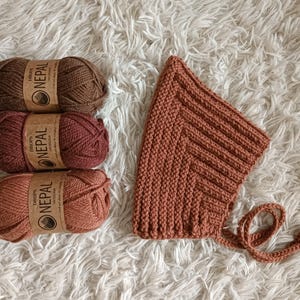 Könnte beinhalten: Draufsicht auf drei Knäuel Drops Nepal-Wolle in Braun- und Rotorangetönen, neben einer gestrickten Mütze in passender Farbe. Die Mütze hat ein strukturiertes Rippendesign und Bindebänder. Das Garn ist mit dem Markennamen versehen.