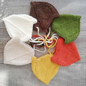 Könnte beinhalten: Sechs gestrickte Baby-Mützen in verschiedenen Farben, darunter Braun, Weiß, Grün, Orange und Gelb. Die Mützen sind in einem Kreis auf einer grauen Holzoberfläche angeordnet.