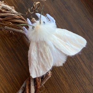 May include: A white moth figurine with a fuzzy, plush-like body and soft, pale wings. The moth features two thin, dark antennae and small, dark eyes, perched upon a brown, twisted twig structure that resembles a natural branch or wreath.