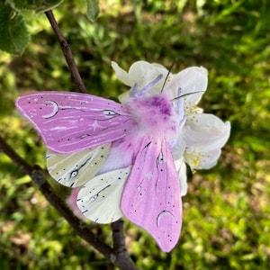 May include: A handcrafted, whimsical moth decoration with lavender wings adorned with crescent moons and stars. The moth has a fluffy purple body and is perched on a white flower with green foliage in the background. The moth's wings are approximately 10 cm wide.
