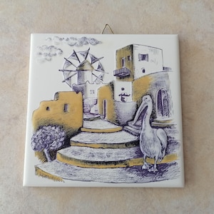 Puede incluir: Un azulejo de cerámica blanco con un diseño pintado a mano de una escena de pueblo griego. El azulejo presenta un molino de viento, edificios, un pelícano y un camino empedrado. El azulejo está pintado en tonos de amarillo, azul y marrón.