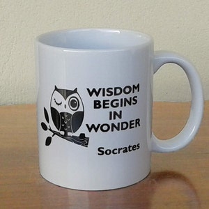 Könnte beinhalten: Weiße Keramiktasse mit einer schwarzen Eulenillustration auf einem Ast. Die Tasse trägt das Zitat "WISDOM BEGINS IN WONDER Socrates" in schwarzem Text. Sie hat einen gebogenen Griff. Die Tasse steht auf einer hellbraunen Oberfläche.