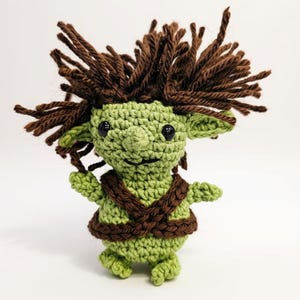 May include: A small, handmade crochet doll featuring a light green body with a textured, yarn-based appearance. The doll has wild, dark brown yarn hair, large black plastic eyes, pointed ears, and a brown yarn harness wrapped around its torso.