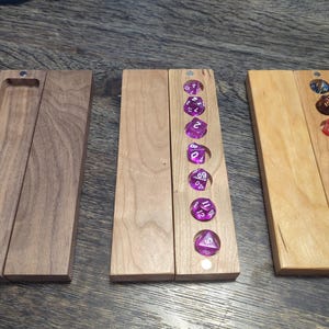 May include: Three rectangular wooden dice cases with magnetic closures. The left case is dark brown walnut, the centre case is light wood containing seven purple translucent dice, and the right case is light wood containing seven multi-coloured dice.