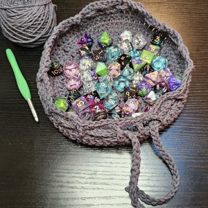 May include: A round, gray crocheted bowl filled with a collection of multi-colored, translucent polyhedral dice. A ball of matching gray yarn and a light green crochet hook are positioned to the left of the bowl, which features a drawstring.