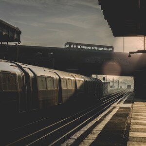 May include: A dark gray train car with windows and doors is parked on a train track. The train is under a bridge and the sun is shining through the clouds.