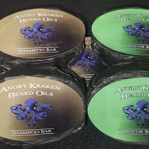 May include: A close-up of six round, black-wrapped bars of soap. The bars are labeled "Angry Kraken Beard Oils" and feature a purple octopus graphic. Two of the bars are labeled "Shampoo Bar" and four are labeled "Pine Tar Soap".