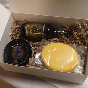 May include: A white gift box filled with brown packing paper. The box contains a small bottle of beard oil, a bar of soap, and a jar of beard oil. The beard oil bottle has a blue label with the text "The Kraken Beard Oil" and a graphic of an octopus. The jar of beard oil has a black label with the text "Angry Kraken Beard Oils" and a graphic of an octopus. The bar of soap is yellow.