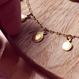 May include: A gold-toned chain necklace featuring multiple small, circular, hammered metal charms suspended at regular intervals along the length of the chain. The charms have a textured, reflective surface and are attached with small jump rings.