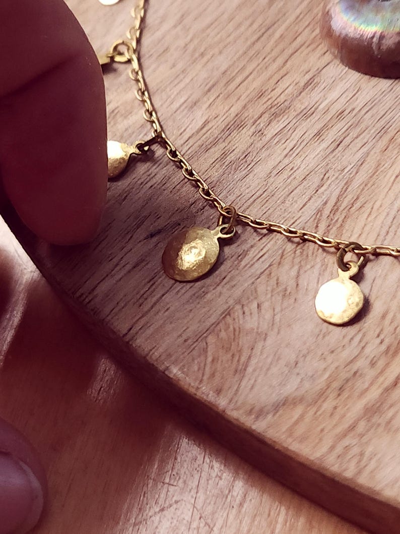 May include: A gold-toned chain necklace featuring multiple small, circular, hammered metal charms suspended at regular intervals along the length of the chain. The charms have a textured, reflective surface and are attached with small jump rings.