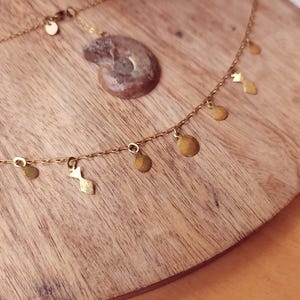 May include: A gold-toned chain necklace featuring small, dangling geometric charms in circular and diamond shapes. The necklace is accompanied by a separate, spiral-shaped fossil pendant with a brownish, textured surface and metallic highlights.