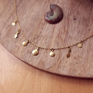 May include: A gold-toned necklace featuring a delicate chain adorned with small, hammered circular and abstract-shaped charms hanging at intervals. The metal components have a textured, matte finish, and the chain includes a small circular tag near the clasp.
