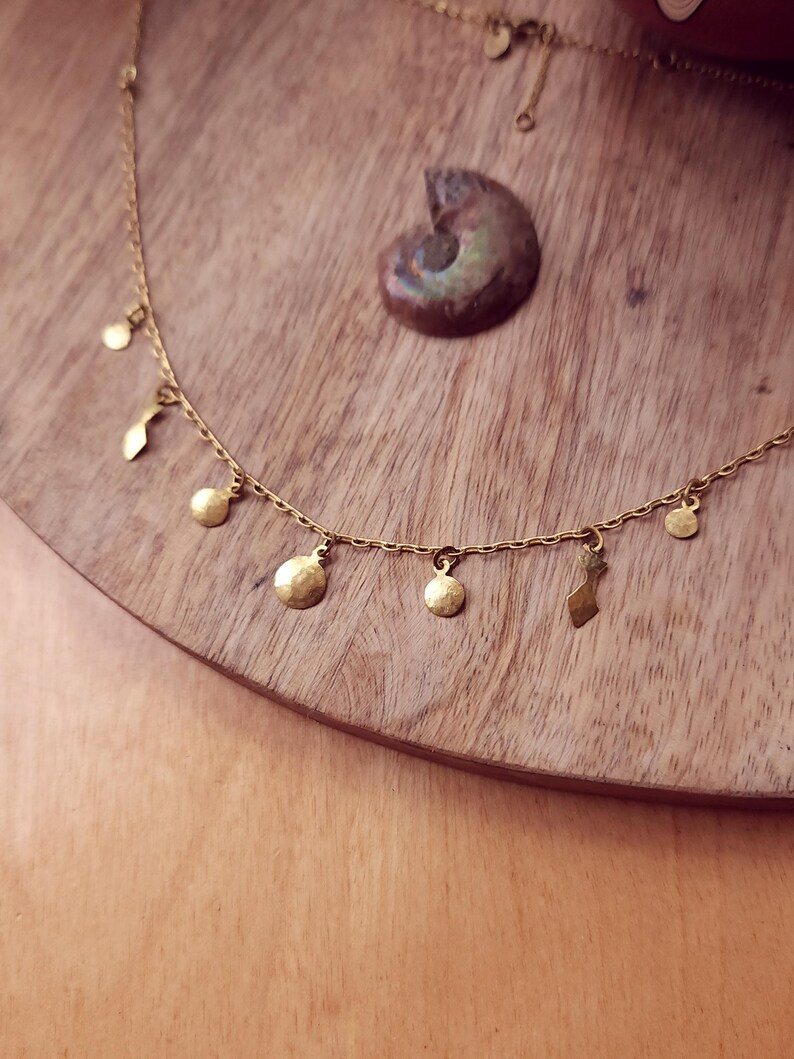 May include: A gold-toned necklace featuring a delicate chain adorned with small, hammered circular and abstract-shaped charms hanging at intervals. The metal components have a textured, matte finish, and the chain includes a small circular tag near the clasp.