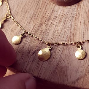 May include: A gold-toned chain necklace featuring multiple small, circular, hammered metal charms suspended at regular intervals along the length of the chain. The charms have a reflective, textured surface and are attached to the chain with small jump rings.