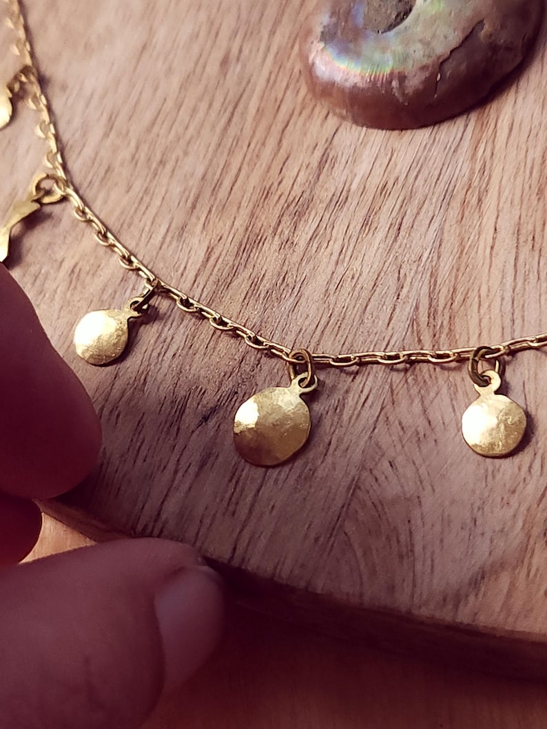 May include: A gold-toned chain necklace featuring multiple small, circular, hammered metal charms suspended at regular intervals along the length of the chain. The charms have a reflective, textured surface and are attached to the chain with small jump rings.
