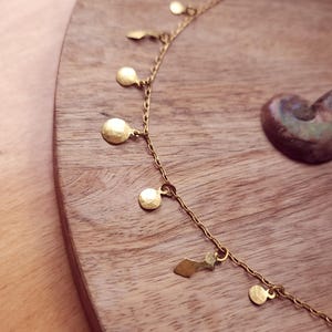 May include: A gold-toned chain necklace featuring a series of small, hammered circular and geometric charms suspended at regular intervals along the length of the delicate metal link chain, creating a layered and textured appearance.