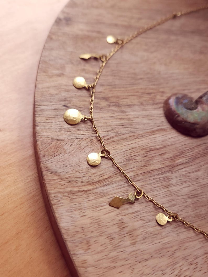 May include: A gold-toned chain necklace featuring a series of small, hammered circular and geometric charms suspended at regular intervals along the length of the delicate metal link chain, creating a layered and textured appearance.