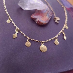 May include: A gold-toned chain necklace featuring a series of small, irregularly shaped, hammered metal charms dangling from the links. The necklace has a delicate, fine-link construction with a clasp closure and a small circular tag near the end.