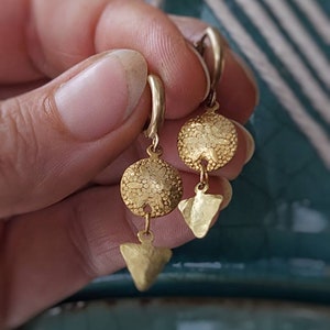 Puede incluir: Un par de pendientes de aro de oro con un dije redondo texturizado y un dije triangular que cuelga de cada aro.