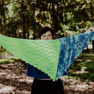May include: A handmade, triangular shawl in shades of green and blue. The shawl is held up, showcasing its knitted texture and color gradient. The green section transitions to a blue section with a textured pattern. The background is a blurred forest.
