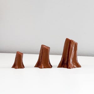 May include: Three brown miniature tree stump figurines of varying heights. The stumps have a textured appearance, with a rough, bark-like surface. They are arranged in a row, showcasing their different sizes.