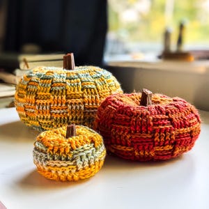 May include: Three knitted pumpkins in different sizes. The largest pumpkin is yellow, orange, and green. The medium pumpkin is red and orange. The smallest pumpkin is yellow, orange, and green. Each pumpkin has a brown stem.