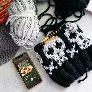 May include: A black and white skull-patterned knitted hat with wooden knitting needles. A ball of gray and white yarn and a gray knitted piece are also visible. A smartphone displays the album art for Dropkick Murphys' "The Walking Dead."