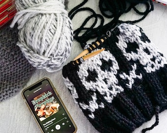 Könnte beinhalten: Eine schwarz-weiß gestrickte Mütze mit Totenkopfmuster und Stricknadeln aus Holz. Ein Knäuel grau-weißer Wolle und ein graues Strickstück sind ebenfalls zu sehen. Ein Smartphone zeigt das Albumcover von Dropkick Murphys' "The Walking Dead".