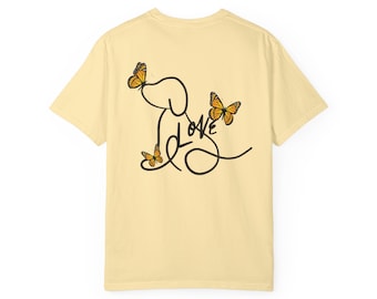 May include: A light yellow t-shirt featuring a black line drawing of a dog with the word "LOVE" incorporated into the design. Three orange butterflies are positioned around the dog. The shirt is made of a soft material.