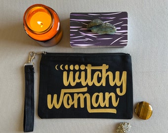 Könnte beinhalten: Schwarze Stofftasche mit goldenem Reißverschluss und goldener Aufschrift "Witchey Woman". Die Tasche liegt neben einer kleinen orangen Kerze, einem kleinen lila und schwarzen Kartenspiel, einem kleinen braunen und goldenen Kristall und einem kleinen silbernen Kristall.
