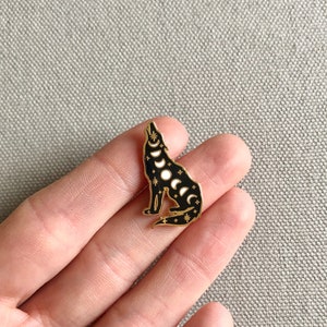 May include: Gold-tone enamel pin featuring a black wolf howling at the moon. The wolf is outlined in gold and has a pattern of stars and moon phases on its body.