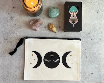 Puede incluir: Una bolsa de lona blanca con una cremallera negra y un diseño de triple luna negro y dorado. La bolsa está sobre una superficie de hormigón gris con una vela encendida, una piedra turquesa, un cristal rosa y una carta de tarot con fondo negro y un diseño geométrico colorido.