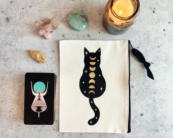 Könnte beinhalten: Eine weiße Canvas-Reißverschlusstasche mit einem schwarzen Katzensilhouetten-Design. Die Katze hat ein goldenes Mondphasen-Design auf dem Rücken. Die Tasche befindet sich auf einer grauen Oberfläche mit einer Tarotkarte, einer Kerze und Kristallen.