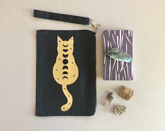 Könnte beinhalten: Schwarze Canvas-Kosmetiktasche mit einer goldenen Katzensilhouette mit Mond- und Sternenmotiv. Die Tasche hat einen schwarzen Riemen.