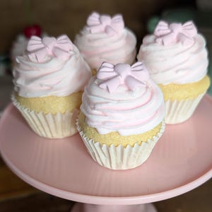 May include: A collection of cupcakes with pink frosting and bow-shaped decorations. The cupcakes are displayed on a pink cake stand. The cupcakes have yellow cake bases and are in white paper liners. Some cupcakes are in the background.