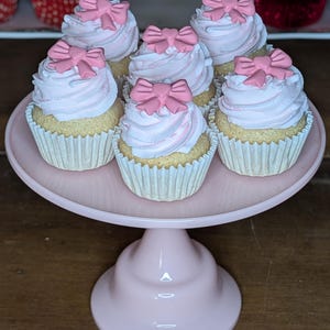 May include: Six cupcakes with pink frosting and pink bow decorations sit on a pink cake stand. The cupcakes have white paper liners and are arranged in a circular pattern. The cake stand is also pink and has a pedestal base.