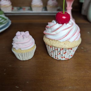 May include: Two decorative cupcakes with white frosting, pink accents, and a red cherry on top. One cupcake is smaller with a pink bow. The cupcakes sit on a wooden surface, with more cupcakes in the background.