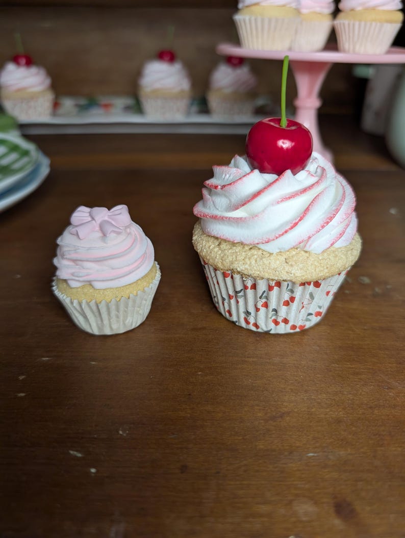 May include: Two decorative cupcakes with white frosting, pink accents, and a red cherry on top. One cupcake is smaller with a pink bow. The cupcakes sit on a wooden surface, with more cupcakes in the background.