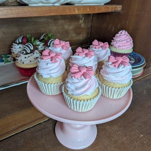 May include: A collection of cupcakes with pink frosting and bow decorations on a pink cake stand. Additional cupcakes with strawberry and chocolate toppings are visible in the background, along with decorative teacups.