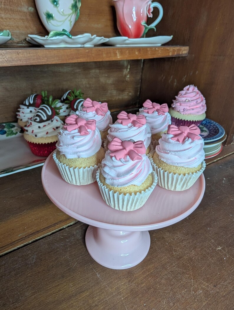 May include: A collection of cupcakes with pink frosting and bow decorations on a pink cake stand. Additional cupcakes with strawberry and chocolate toppings are visible in the background, along with decorative teacups.