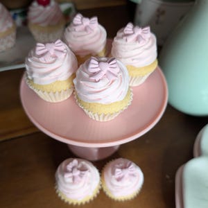 May include: A collection of cupcakes with pink frosting and bow-shaped decorations. The cupcakes are displayed on a pink cake stand and a white plate. The cupcakes have a light yellow base.