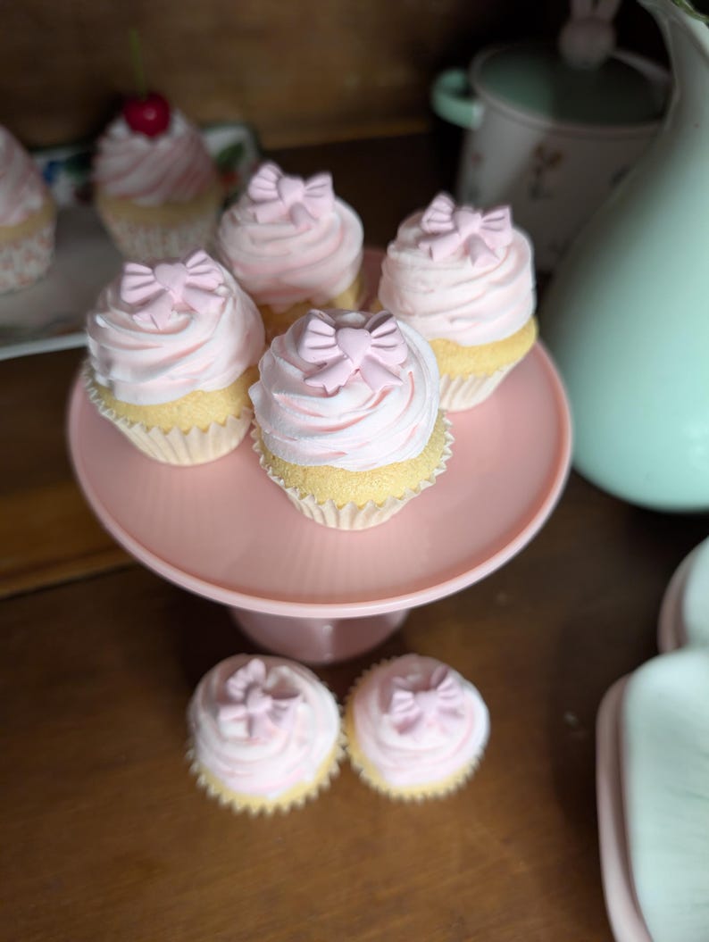 May include: A collection of cupcakes with pink frosting and bow-shaped decorations. The cupcakes are displayed on a pink cake stand and a white plate. The cupcakes have a light yellow base.