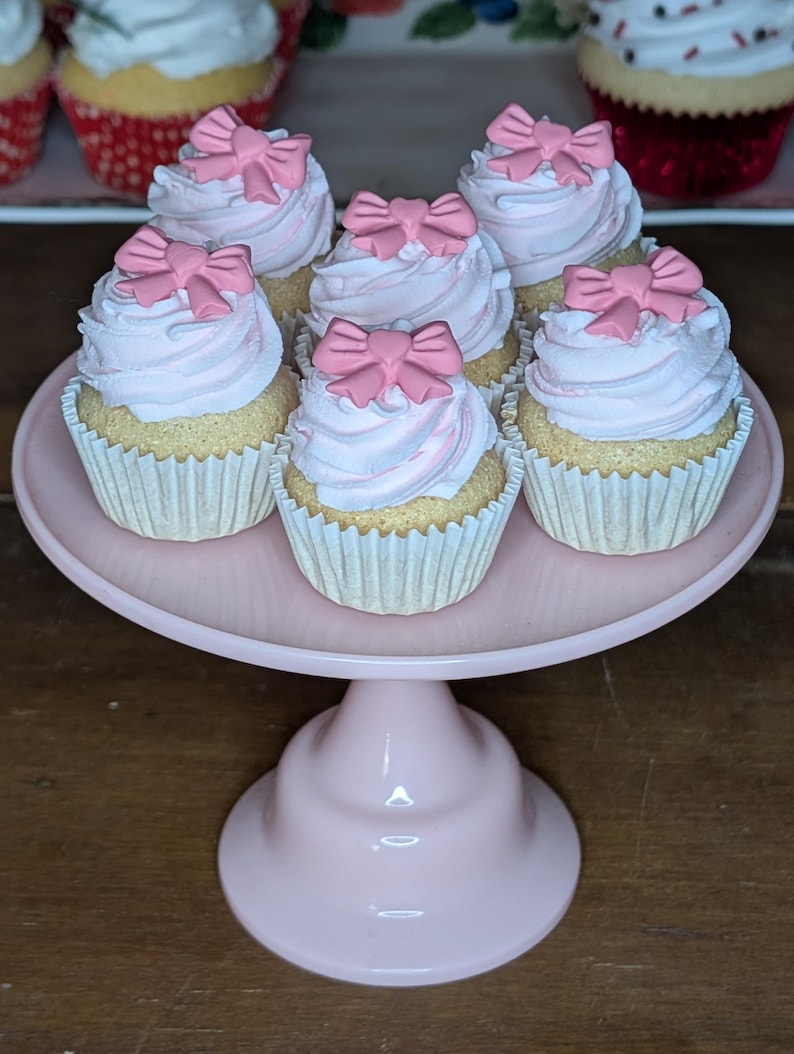 May include: Six cupcakes with pink frosting and pink bow decorations sit on a pink cake stand. The cupcakes have white paper liners and are arranged in a circular pattern. The cake stand is also pink and has a pedestal base.