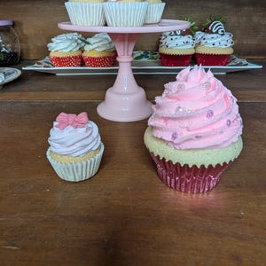 May include: A collection of decorative cupcakes with pink frosting and various toppings. Some cupcakes are displayed on a pink tiered stand, while others are arranged on a wooden surface. The cupcakes have white paper liners.