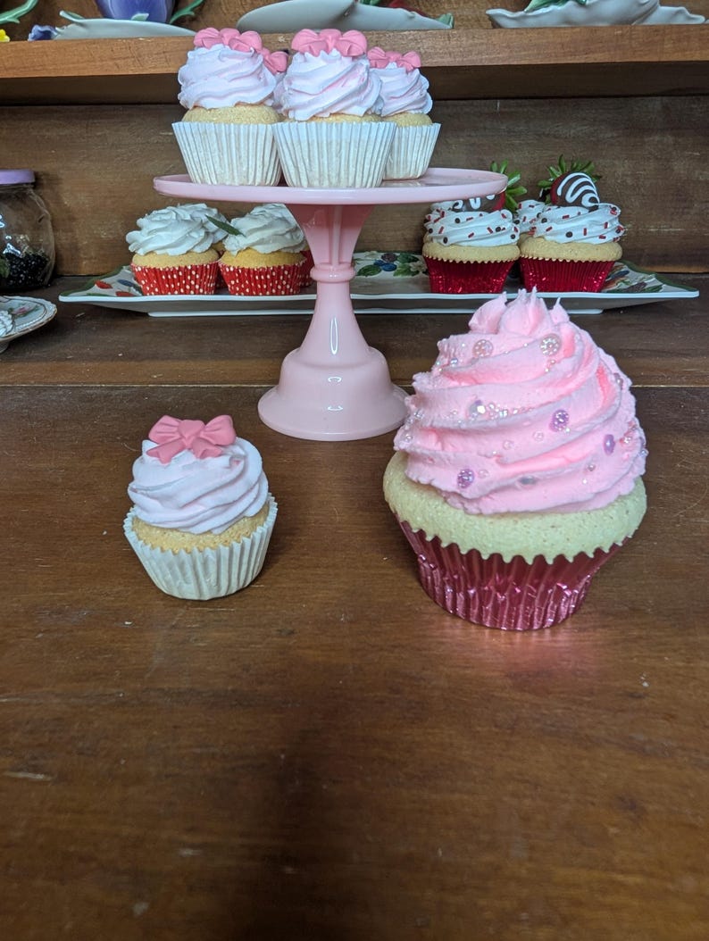 May include: A collection of decorative cupcakes with pink frosting and various toppings. Some cupcakes are displayed on a pink tiered stand, while others are arranged on a wooden surface. The cupcakes have white paper liners.