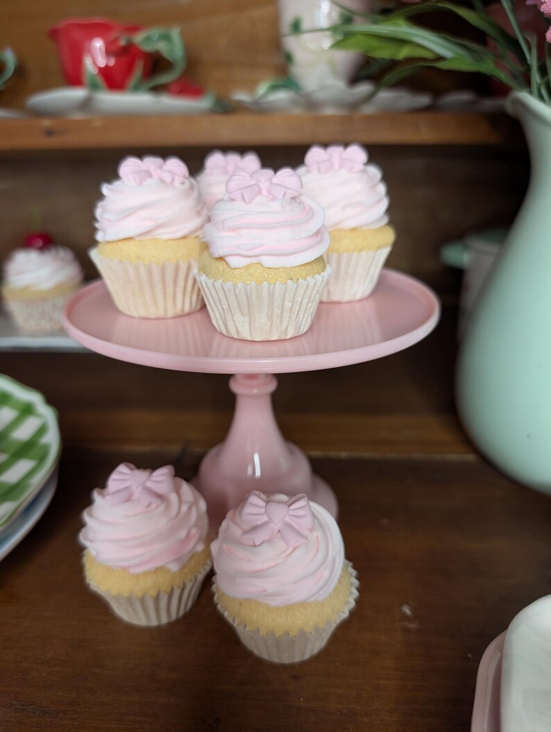 May include: A collection of cupcakes with pink frosting and bow-shaped decorations. The cupcakes are displayed on a pink cake stand and are set against a wooden background. The cupcakes have a light yellow base.