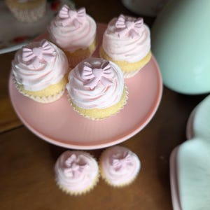 May include: A collection of cupcakes with pink frosting and bow-shaped decorations. The cupcakes are arranged on a pink cake stand and a floral plate. The image has a soft, pastel color palette, suggesting a celebratory or dessert theme.