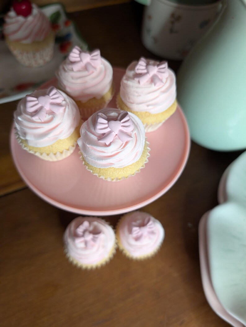 May include: A collection of cupcakes with pink frosting and bow-shaped decorations. The cupcakes are arranged on a pink cake stand and a floral plate. The image has a soft, pastel color palette, suggesting a celebratory or dessert theme.