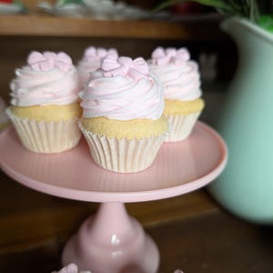 May include: Pink frosted cupcakes with bow decorations on a pink cake stand. The cupcakes have yellow cake bases and are in white paper liners. A light green vase with flowers is in the background.